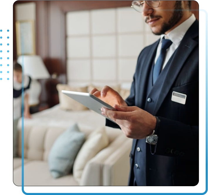 Young elegant manager of luxurious hotel using tablet against chambermaid changing bedclothes and preparing room for new guests